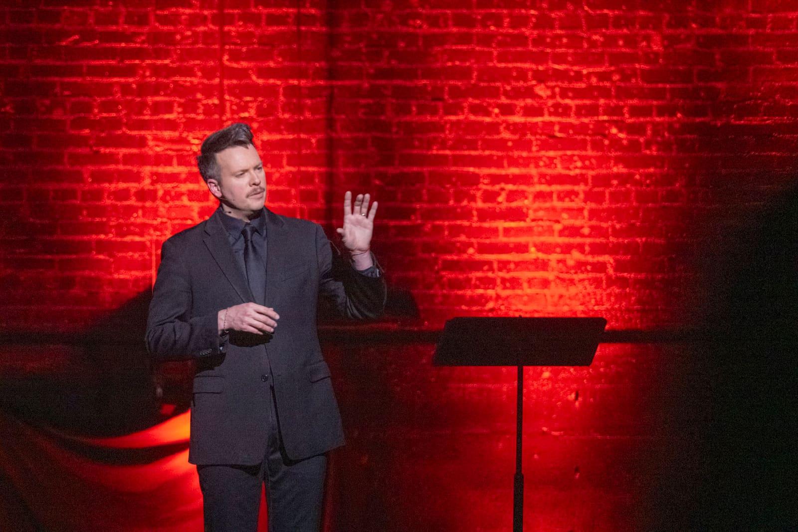 An adult gestures while speaking on a red-lit stage.