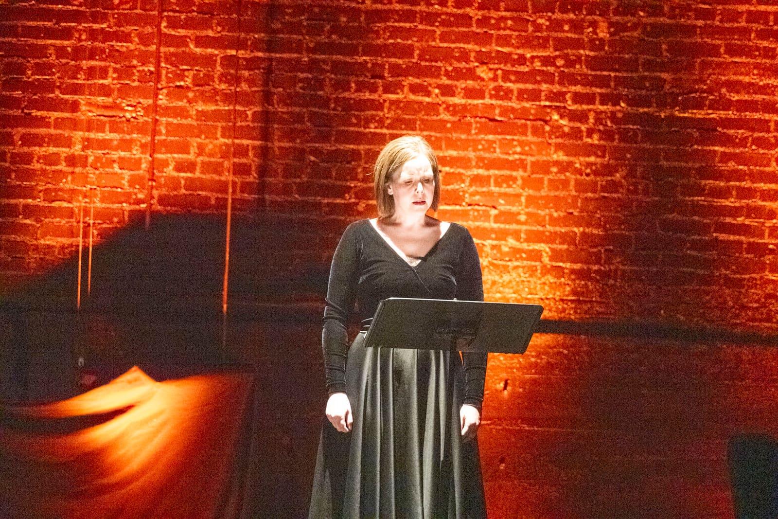 An adult sings at a music stand against a red-lit brick wall.