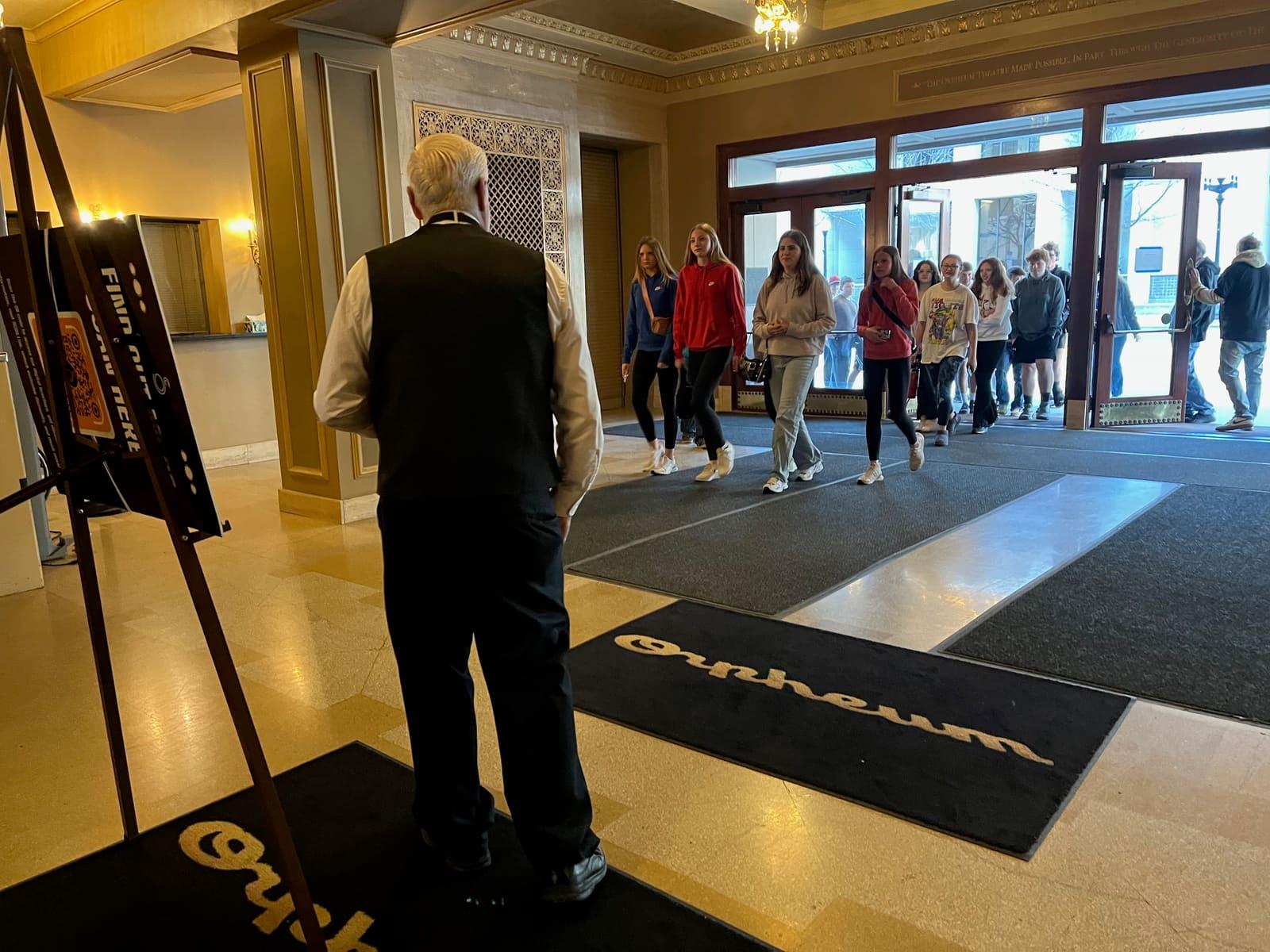 An adult stands in a lobby while students line up near the entrance.
