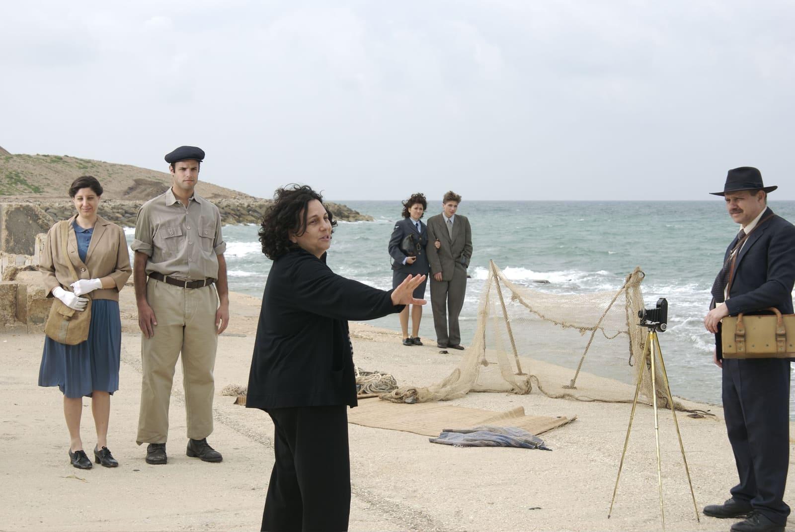 Costumed adults stand on a beach as a woman gestures between two uniformed men.