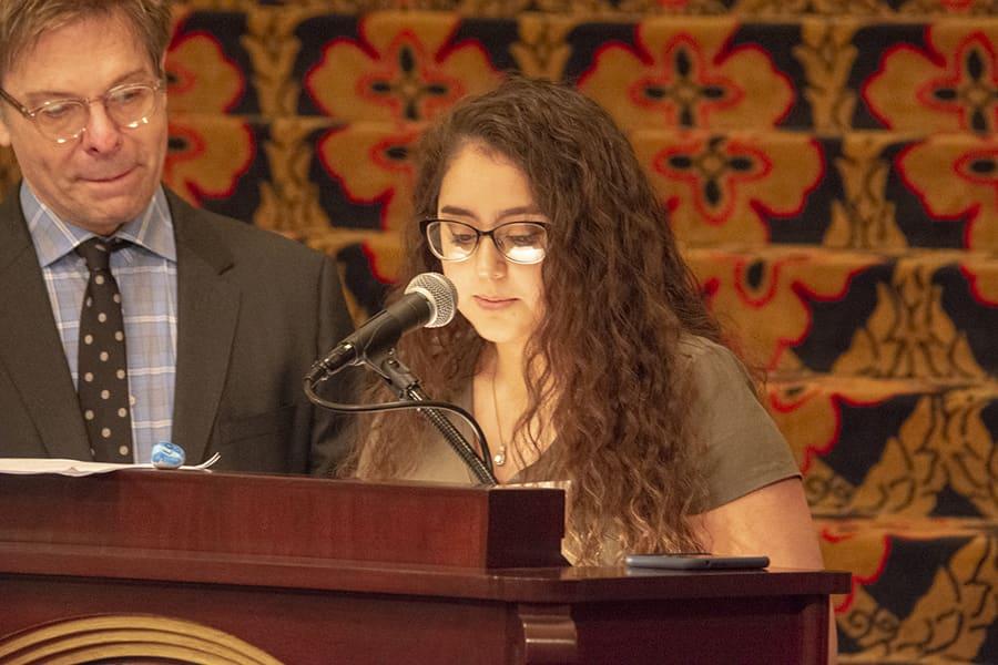 A young woman reads at a lectern microphone while a man stands beside her.