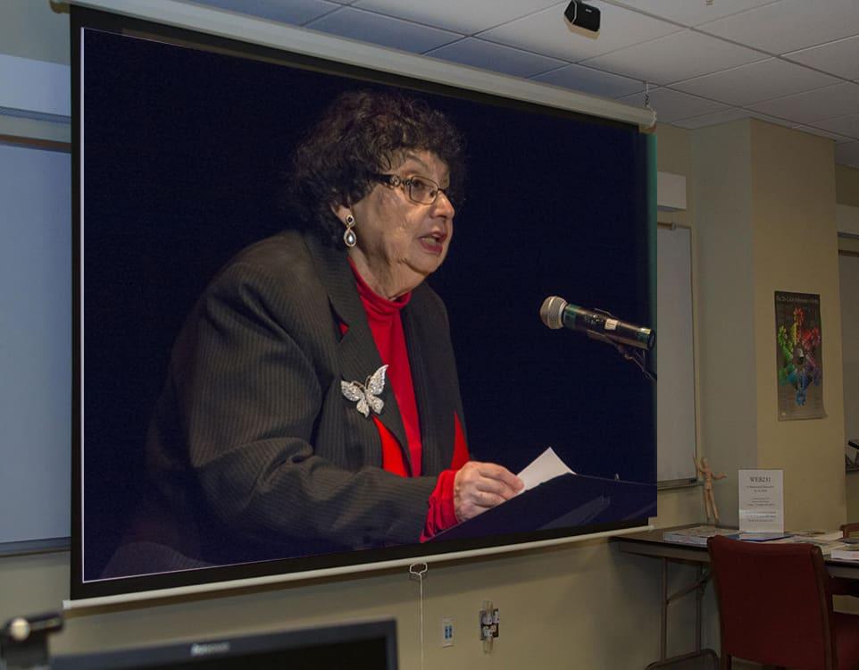 A classroom projection shows an older woman speaking at a microphone.