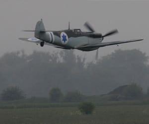 A dark propeller airplane flies low over a grassy field beneath a gray sky.