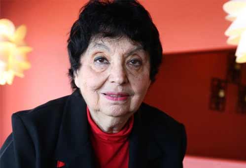 Portrait of an older woman in a black jacket and red shirt against a red wall.