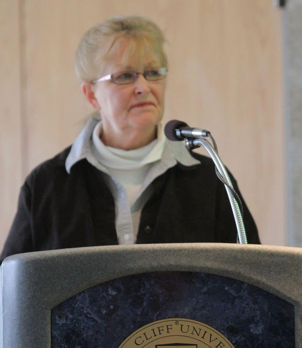 A woman with short blond hair speaks at a podium microphone.