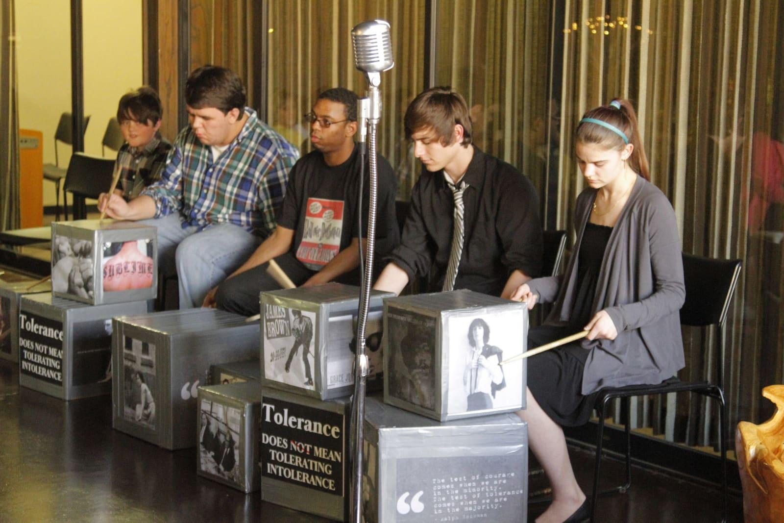 Five people sit behind hand drums and microphones in a curtained room.