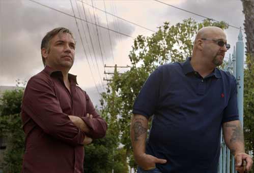 Two men stand outdoors beside a chain-link fence under a cloudy sky.