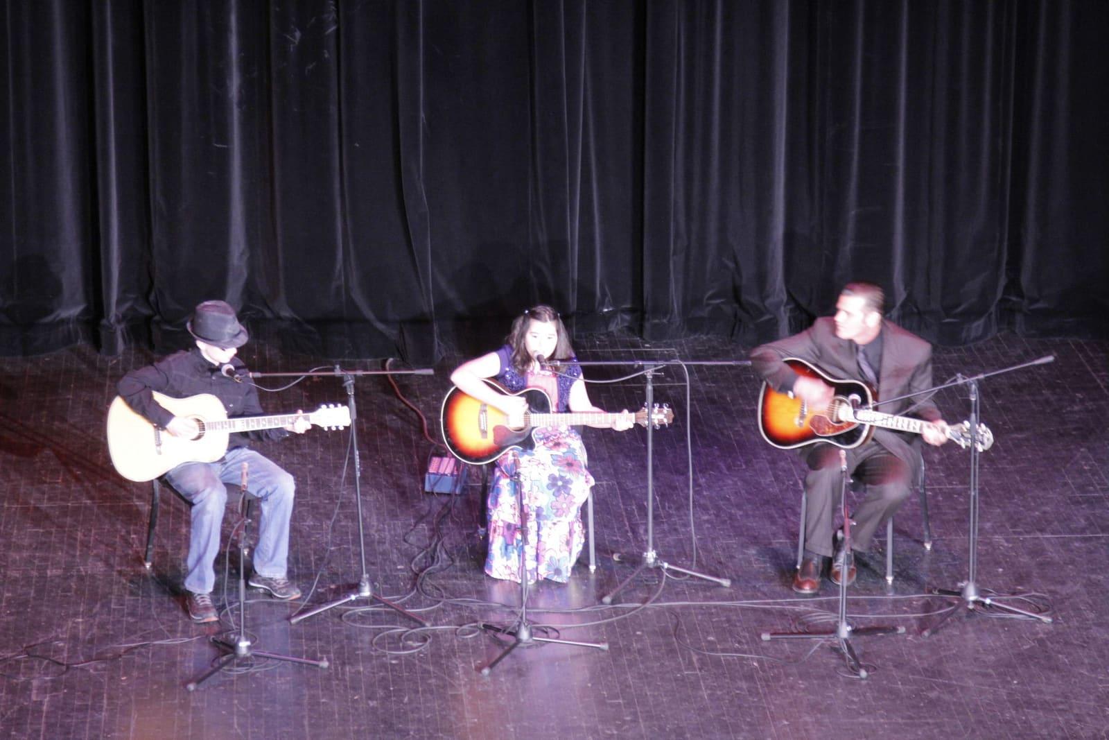Three musicians sit onstage playing acoustic guitars at microphones.