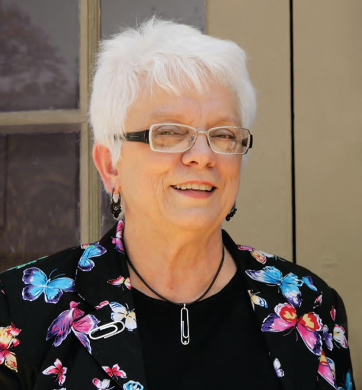 Portrait of an older woman with white hair and glasses wearing a black floral jacket.
