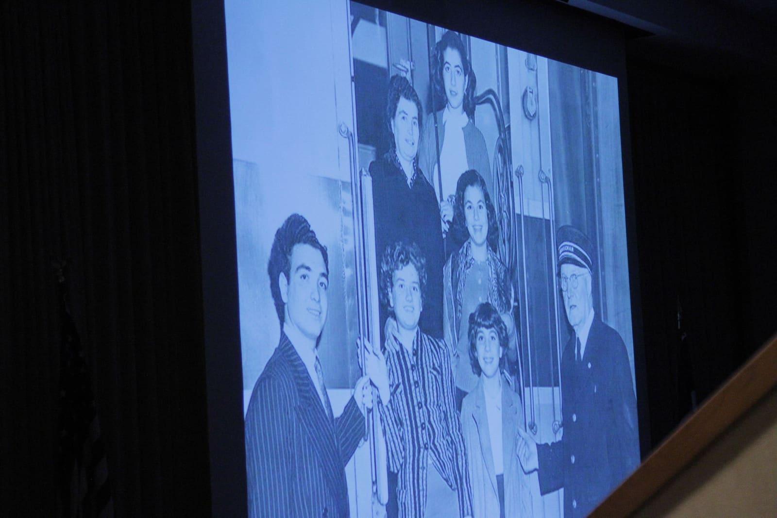 A projection screen displays a black-and-white family photograph in a dark auditorium.