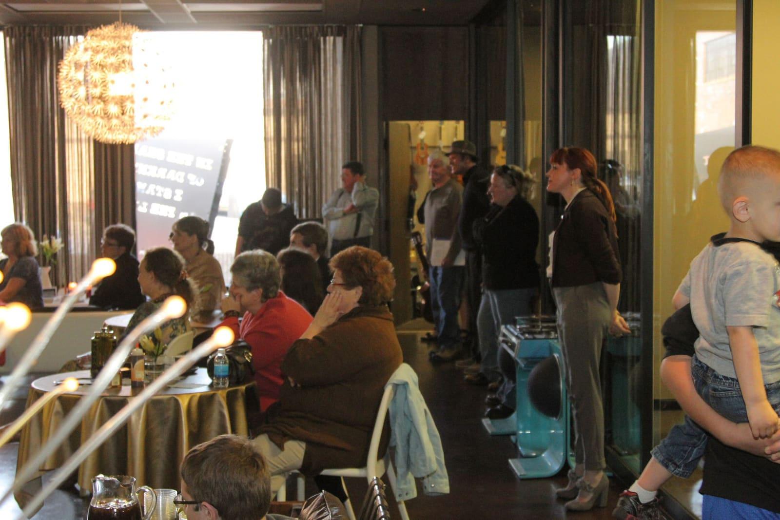 People sit and stand in a crowded room with white taper candles in the foreground.