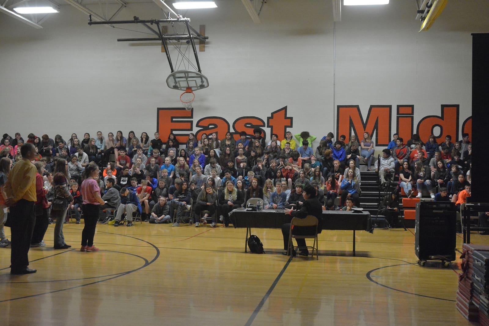 A large audience fills gymnasium bleachers behind speakers and music equipment on the floor.