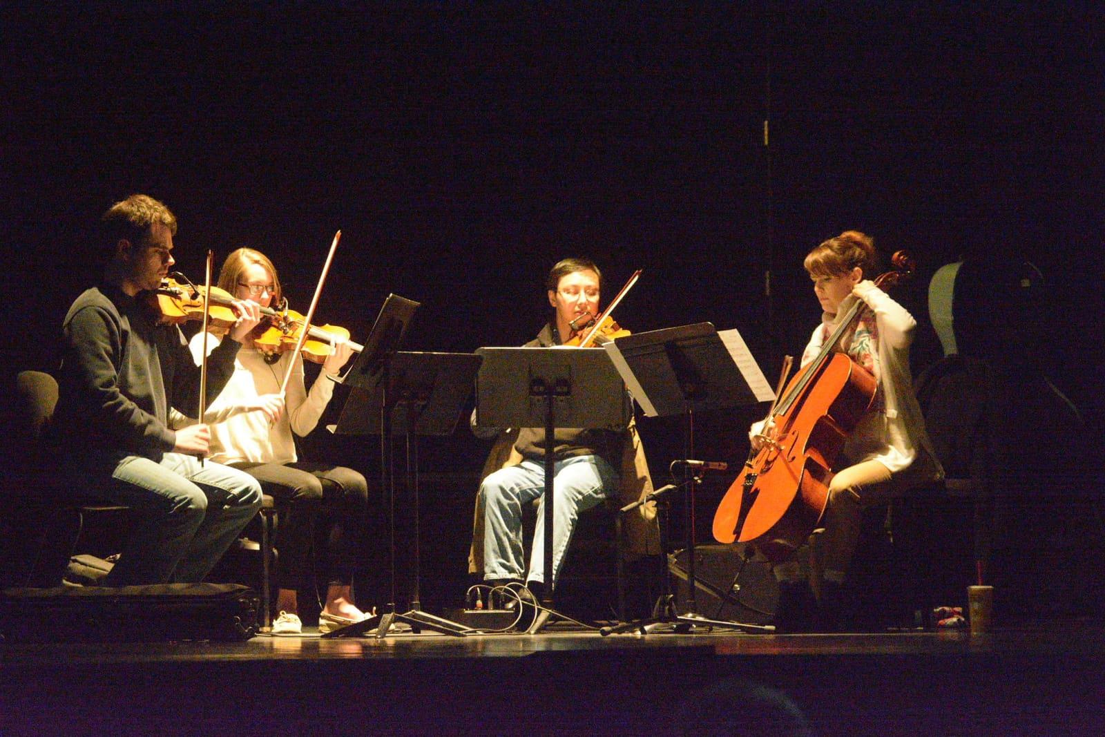 Four seated string musicians perform under spotlights on a dark stage.