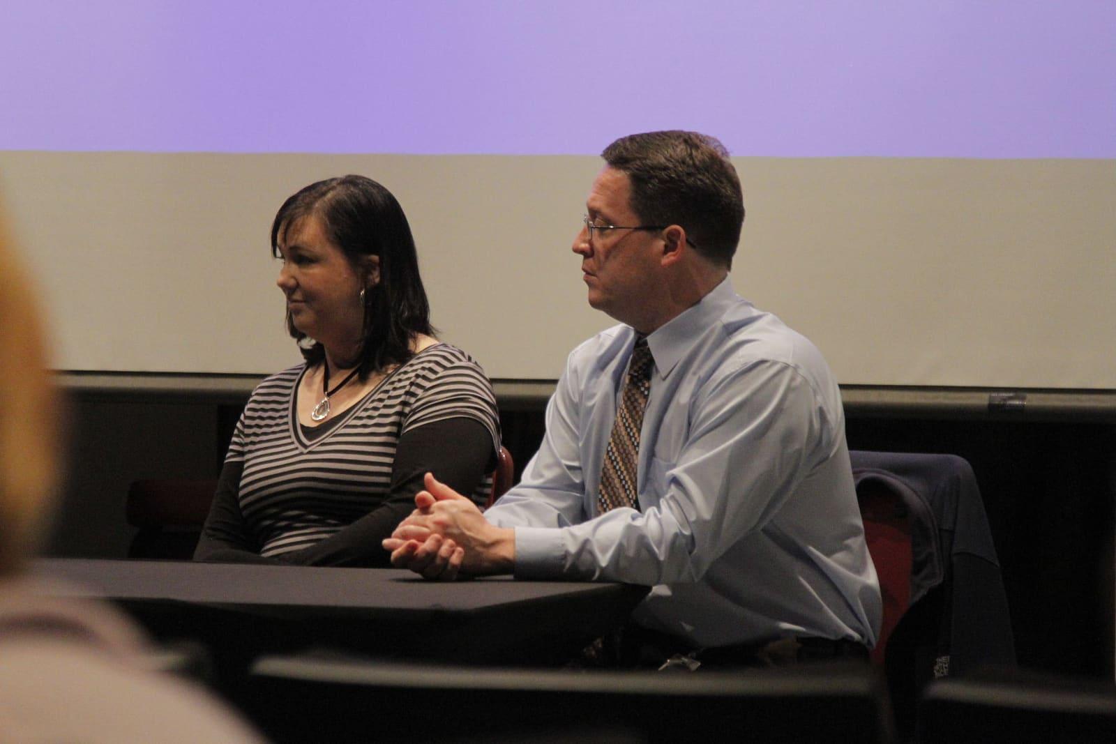 A woman and man sit side by side at a table during a public discussion.