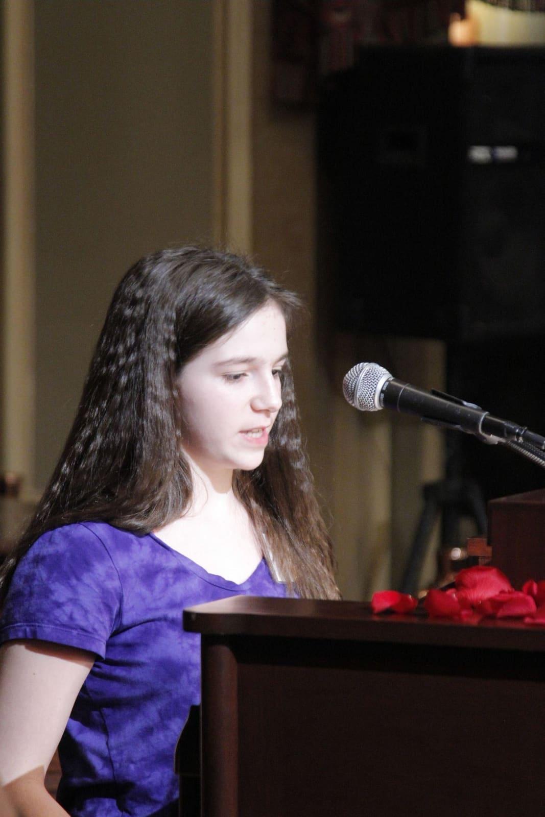 A young woman reads at a microphone behind a wooden lectern.